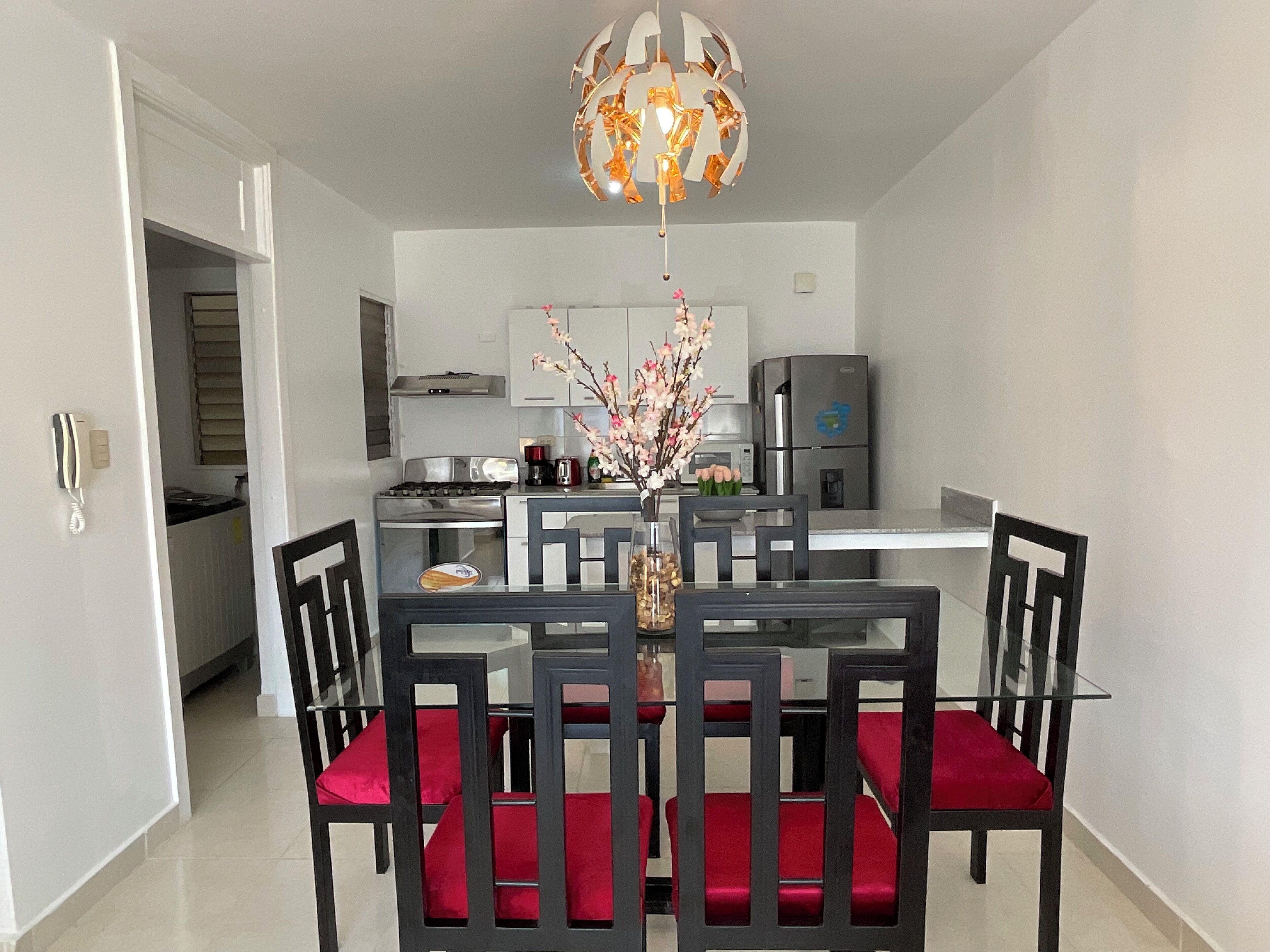 Une cuisine moderne dotée d’appareils électroménagers en acier inoxydable, un coin repas avec une table en verre et des chaises à coussins rouges, ainsi qu’un lustre décoratif.
