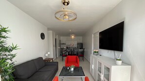 Ein modernes Wohnzimmer mit einer schwarzen Couch, roten Sesseln, einem Glastisch, einem Fernseher auf einer weißen Kommode und einem Essbereich im Hintergrund.