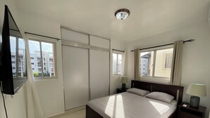 Ein Schlafzimmer mit einem Bett, zwei Nachttischen, einem an der Wand befestigten Fernseher und großen Fenstern mit Vorhängen.