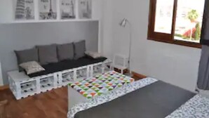 A small, neatly arranged room with a bed, a bench with cushions, a desk, and a chair. There are framed pictures on the wall and a window with a view of the outside.