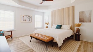 A bedroom with a large bed, a wooden bench, a ceiling fan, and a window with blinds. - 30A Oasis with Lake, Beach, Patio, Fenced Yard for Pets (Panama City Beach)