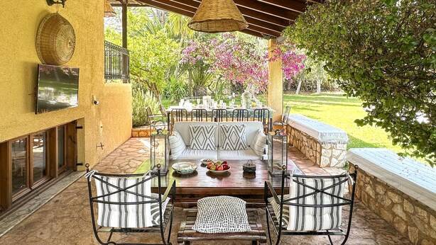 Un porche cubierto con una mesa de comedor para cuatro personas, rodeado de sillas de mimbre y un sofá con cojines estampados.