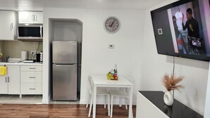 A modern kitchen with white cabinets, a stainless steel refrigerator, and a microwave. There's a dining table with chairs and a small decorative vase. A clock is on the wall, and a TV is mounted above it. - Studio Room in Halifax (Halifax)