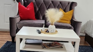 A living room with a dark blue rug, a white coffee table, a dark brown sofa with red and yellow pillows, and a framed picture on the wall. - Studio Room in Halifax (Halifax)