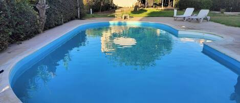 Una piscina con borde curvo, rodeada de un solárium empedrado. Hay sillones de descanso y una zona cubierta con mesas y sillas.