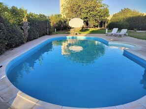 A swimming pool with a curved edge, surrounded by a paved deck. There are lounge chairs and a covered area with tables and chairs. - Όμορφο εξοχικό στούντιο με πισίνα & κήπο,εύκολη πρόσβαση, 6χλμ από αεροδρόμιο (Kokkini Hani)