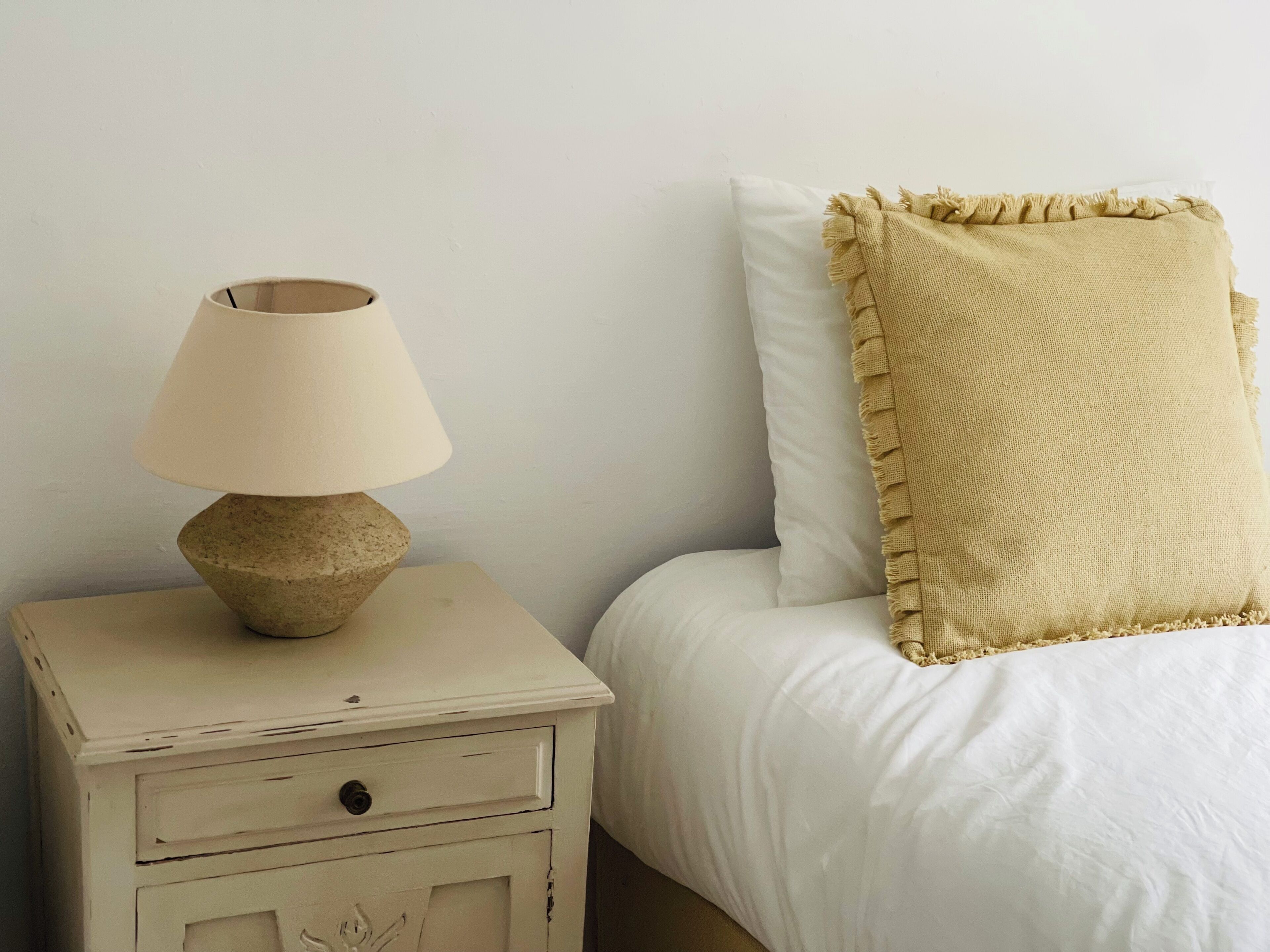 Una mesita de noche pequeña con una lámpara, un almohadón blanco y una cama con una manta texturizada.