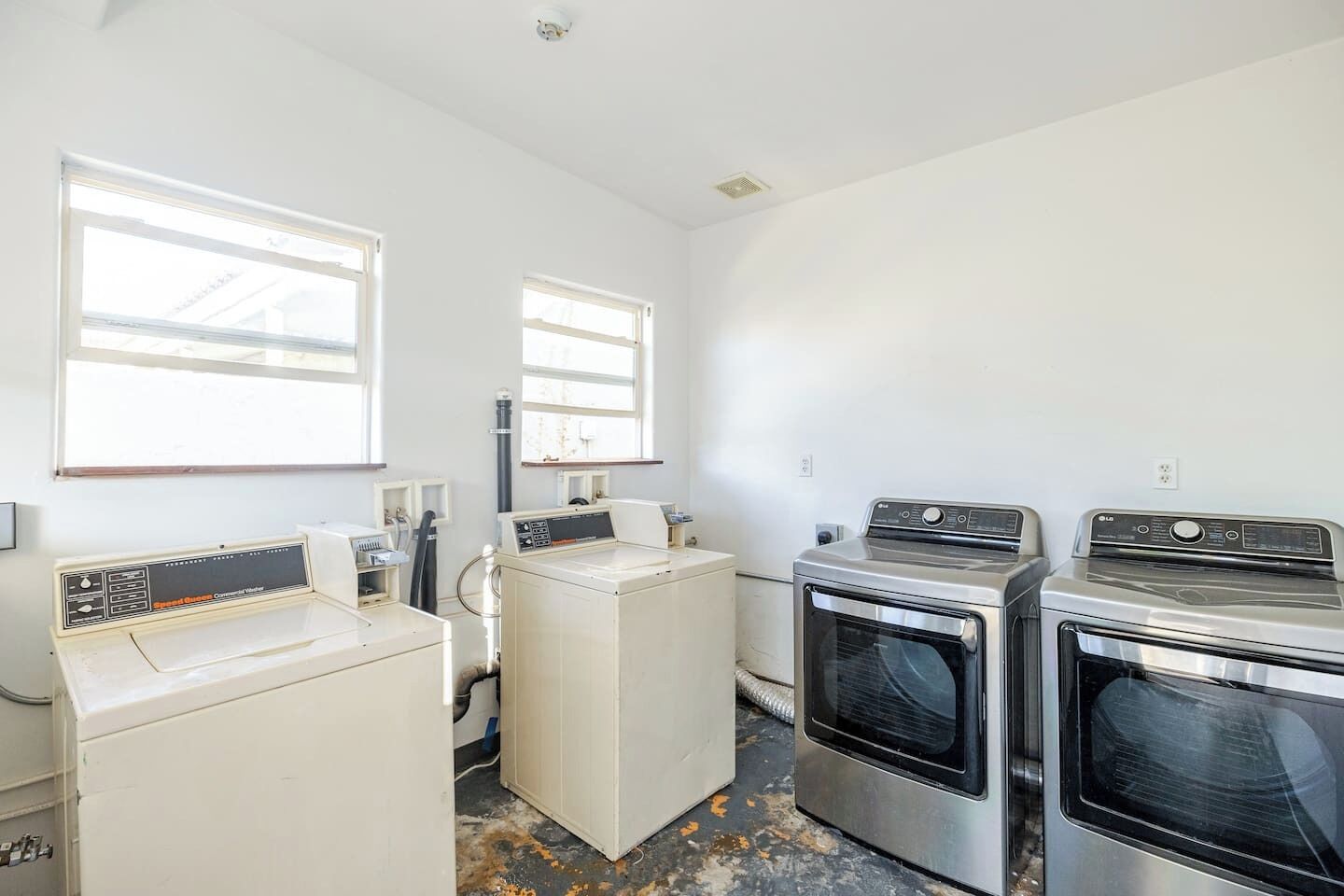 A laundry room with three washing machines and two dryers, white walls, and large windows.
