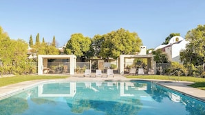 Une piscine dotée de chaises longues et d’un espace de repos couvert, entourée d’arbres et bordée d’un bâtiment blanc surmonté d’un toit en forme de dôme.