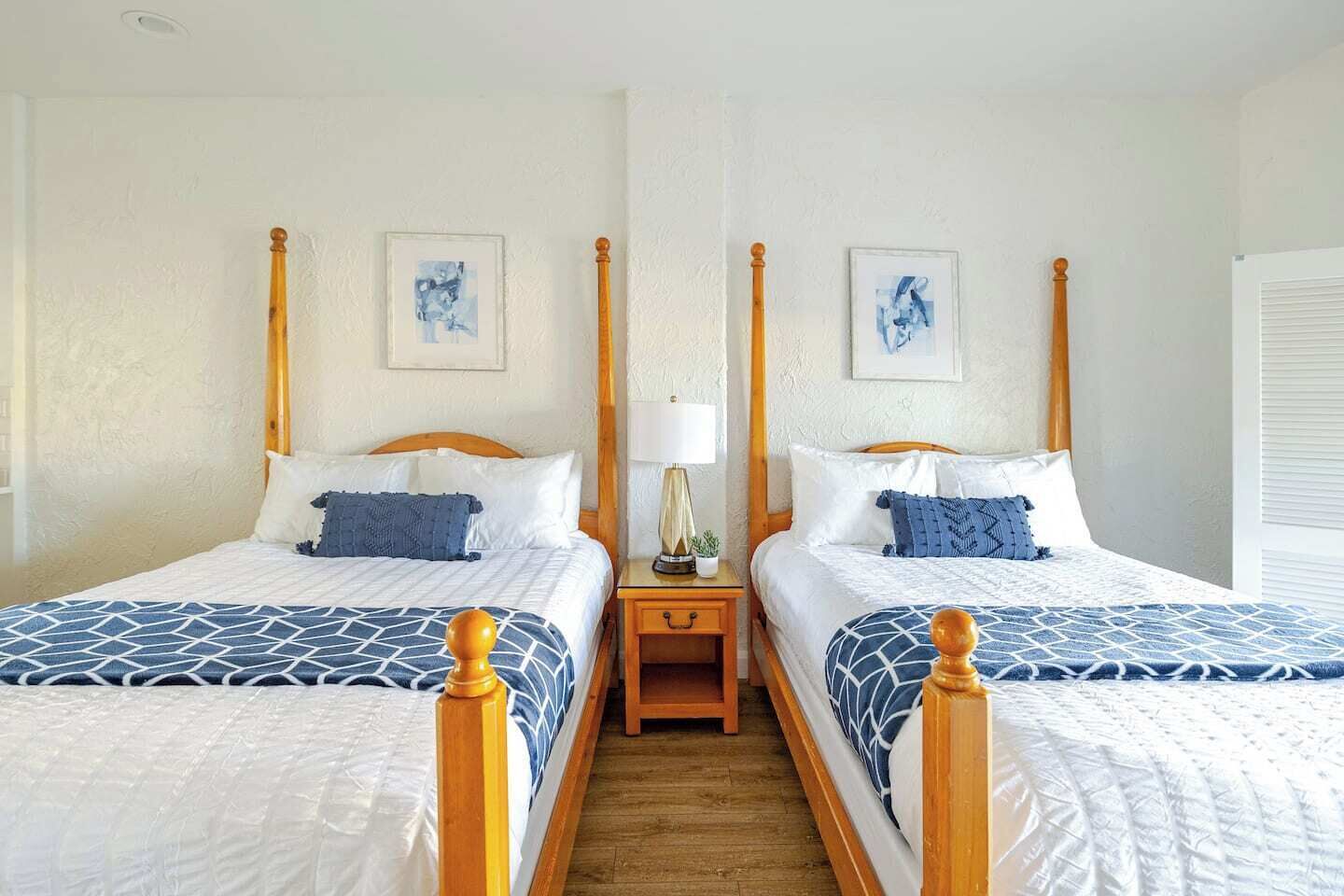 Een tweepersoonskamer met twee bedden, elk met een houten hoofdbord, wit beddengoed en blauwe bedspreien. Tussen de bedden staat een nachtkastje met een lamp en een kleine plant.