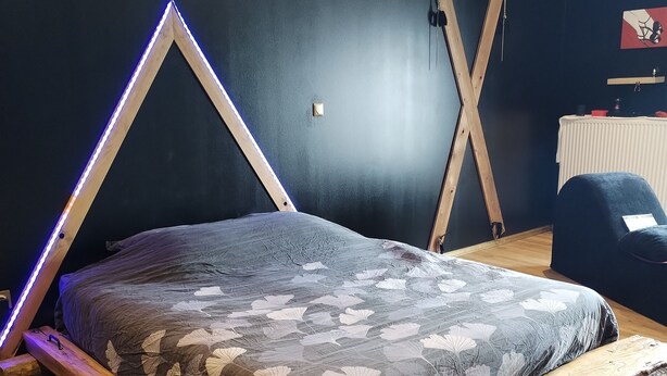 Un llit amb estructura de fusta i un llençol amb motius de fulles en una habitació amb parets fosques i un sòl de fusta.