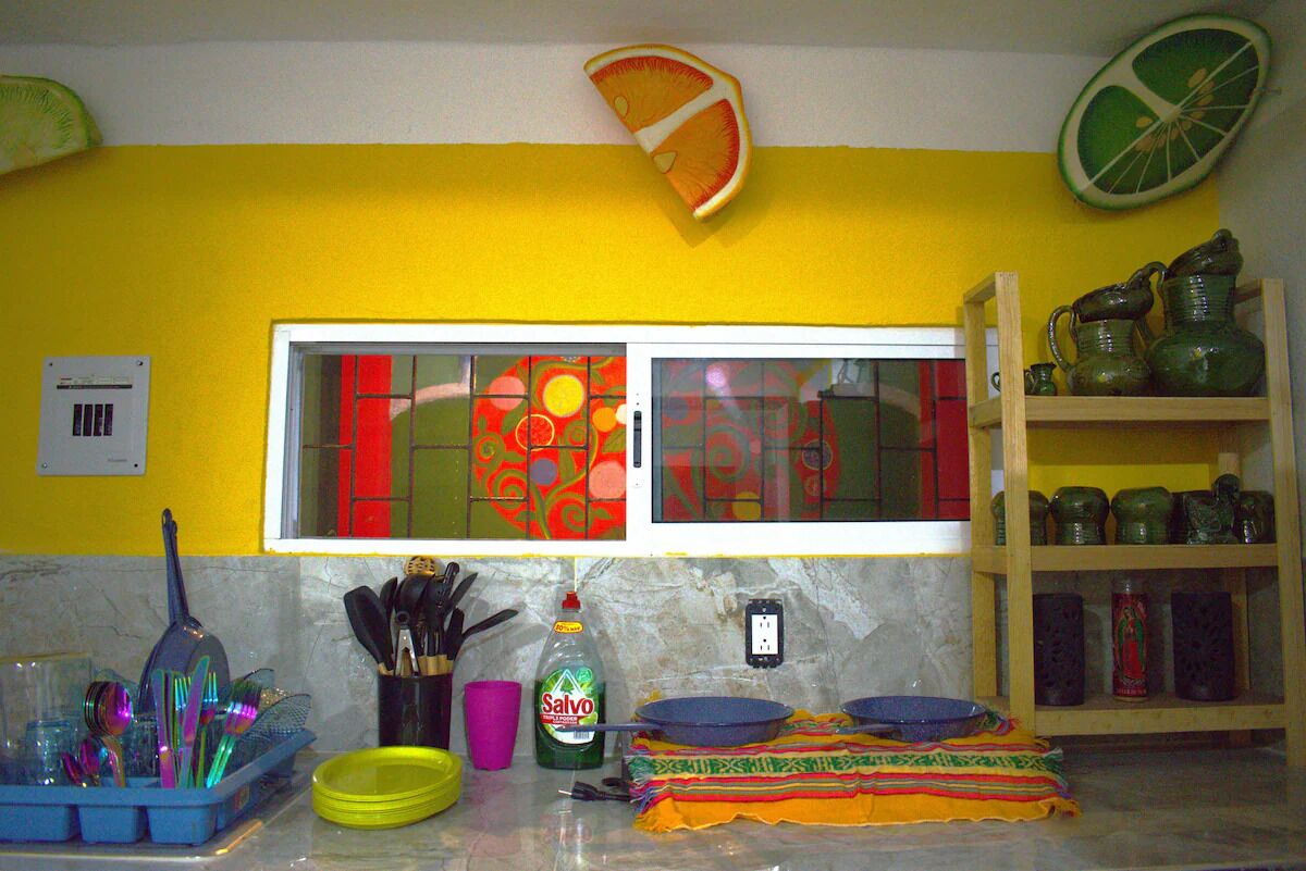 A kitchen with a yellow wall, a window with red and orange patterns, and a shelf with green jars.