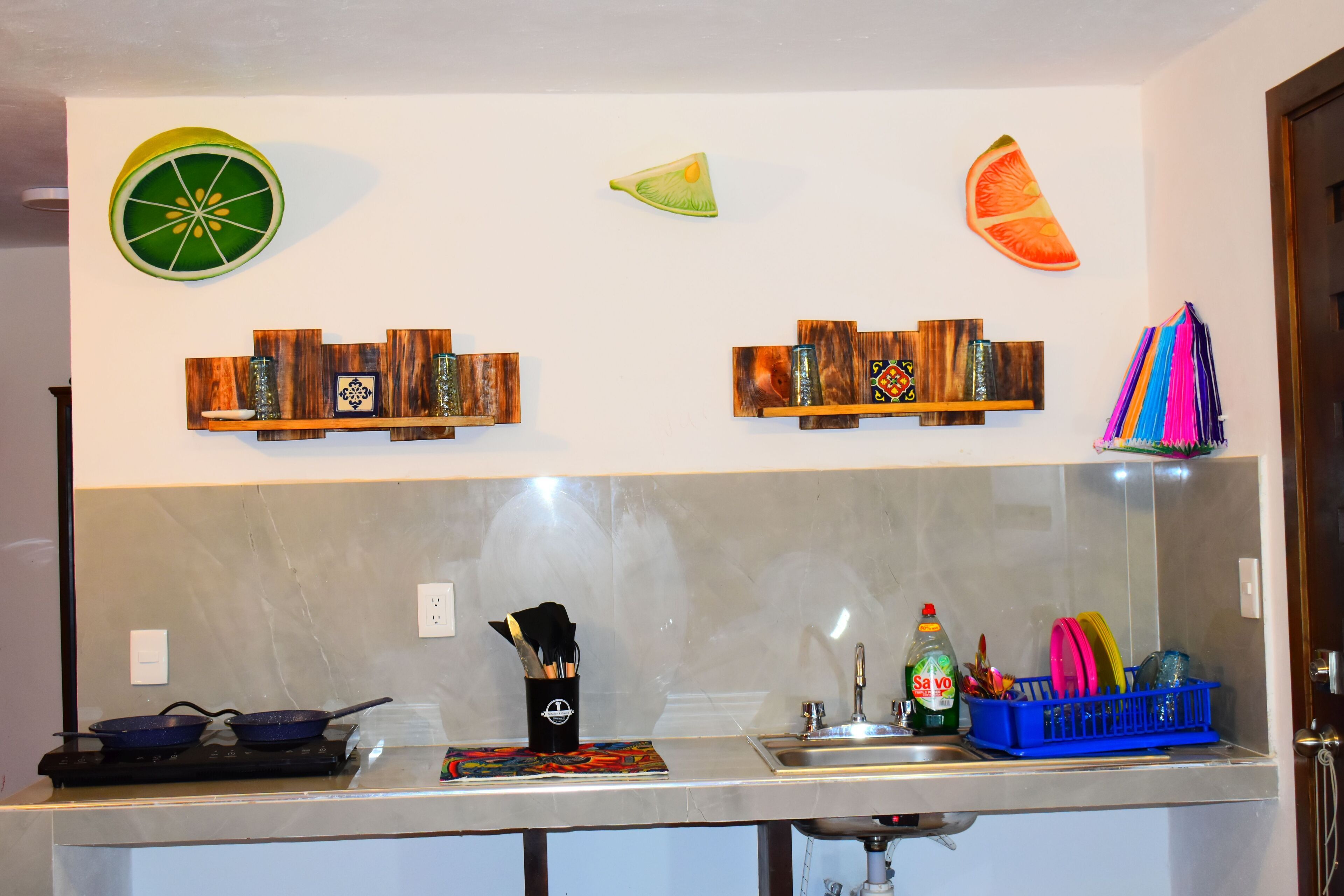 Una cocina con estufa, lavamanos y varios utensilios de cocina. Se observan elementos decorativos en la pared y un colorido tejido.