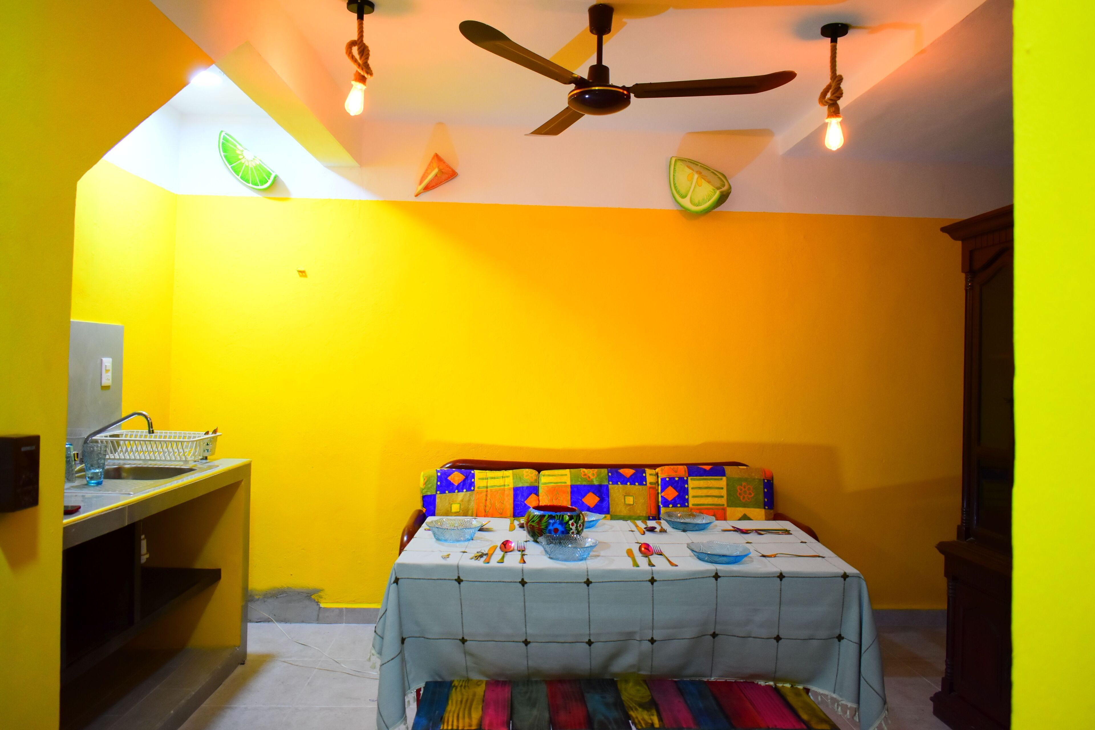 A room with a bed, a table with a tablecloth, a ceiling fan, and a light fixture.