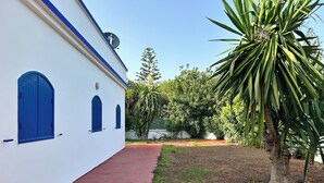 Un bâtiment blanc avec des portes et des fenêtres bleues, un chemin rouge et une végétation luxuriante tout autour.