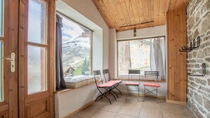 A room with wooden walls and a stone wall, a window with a view of a mountain, a table with chairs, and a hanging light fixture. - LES FLOCONS - BONNEVAL SUR ARC (BONNEVAL SUR ARC)