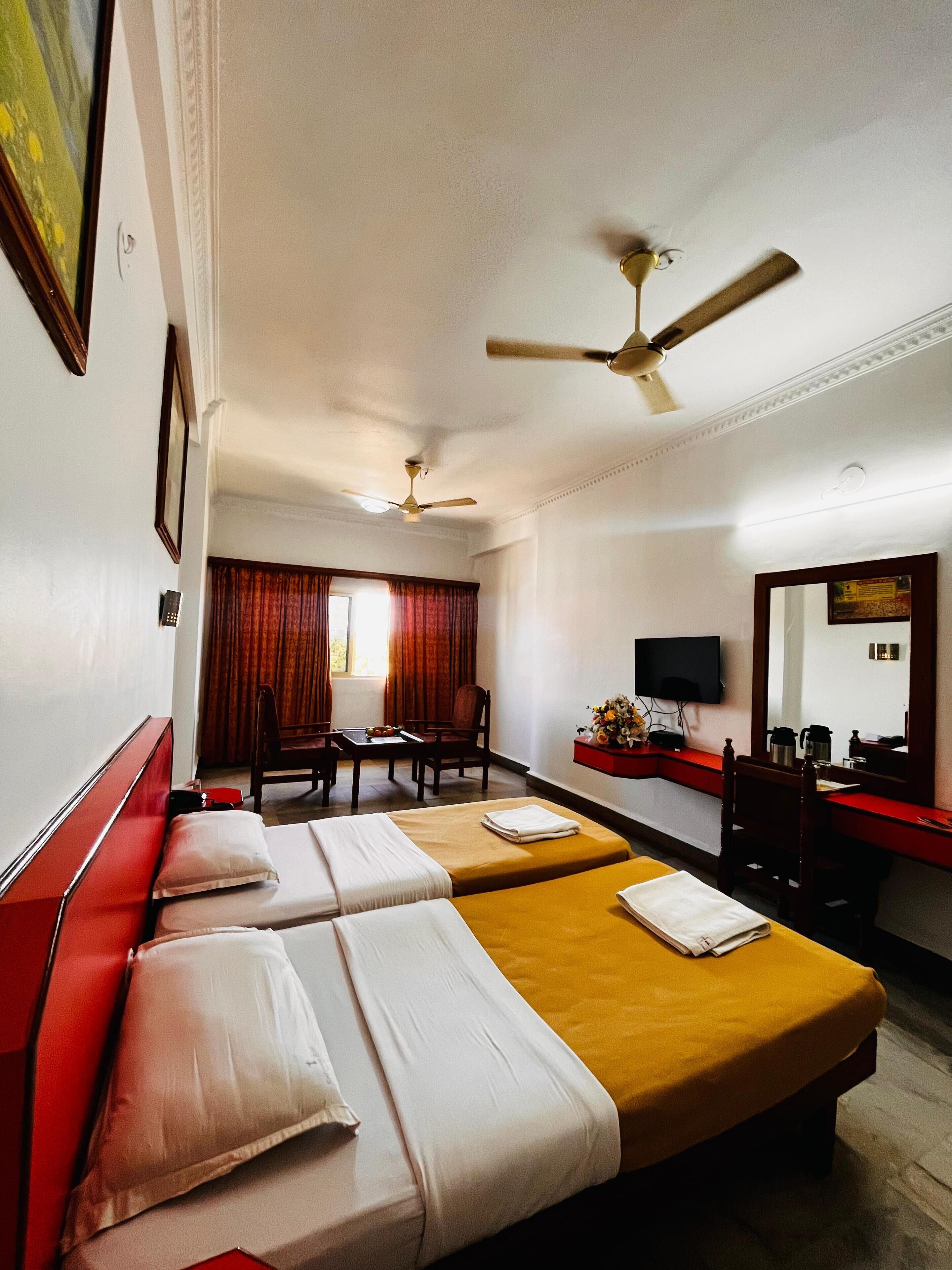 A hotel room with two beds, a desk, a chair, a television, and a ceiling fan.