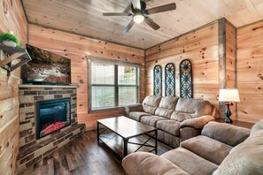 A cozy living room with a fireplace, wooden walls, a ceiling fan, and a large window with blinds. - Free Dollywood Tix/10Min2Dollywood/DogsOK (Sevierville)
