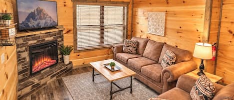 A cozy cabin interior with a fireplace, a television, a sofa, and a window with blinds.