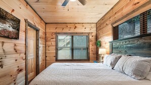 A wooden cabin bedroom with a bed, a window with blinds, a ceiling fan, and a wall-mounted lamp.