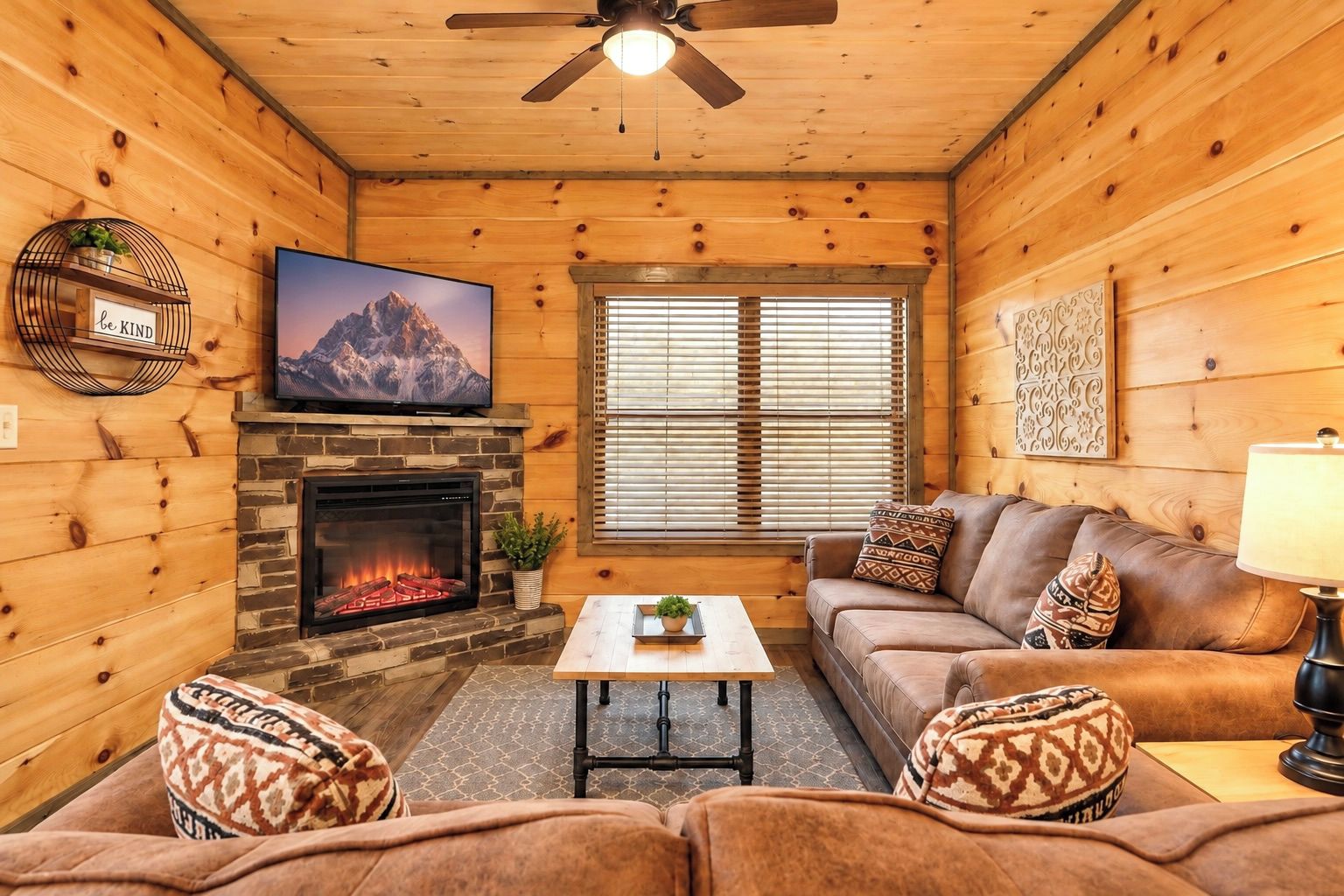 A cozy cabin interior with a fireplace, a television mounted above it, a sofa set, and a window with blinds.