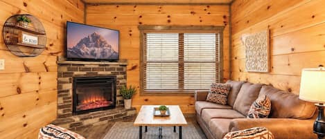 Interior acogedor de una cabaña con chimenea, un televisor montado encima, un conjunto de sofás y una ventana con persianas.