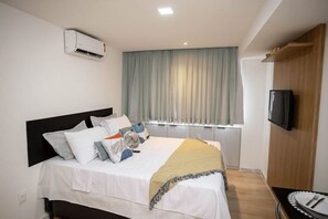 Ein ordentlich bezogenes Bett mit weißer Decke und mehreren Kissen, eine fest installierte Klimaanlage, ein an der Wand befestigter Fernseher und ein Fenster mit hellblauen Vorhängen.