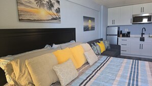Un dormitorio con cama, mesita de noche, un cuadro de un atardecer, kitchenette con microondas y lavabo.