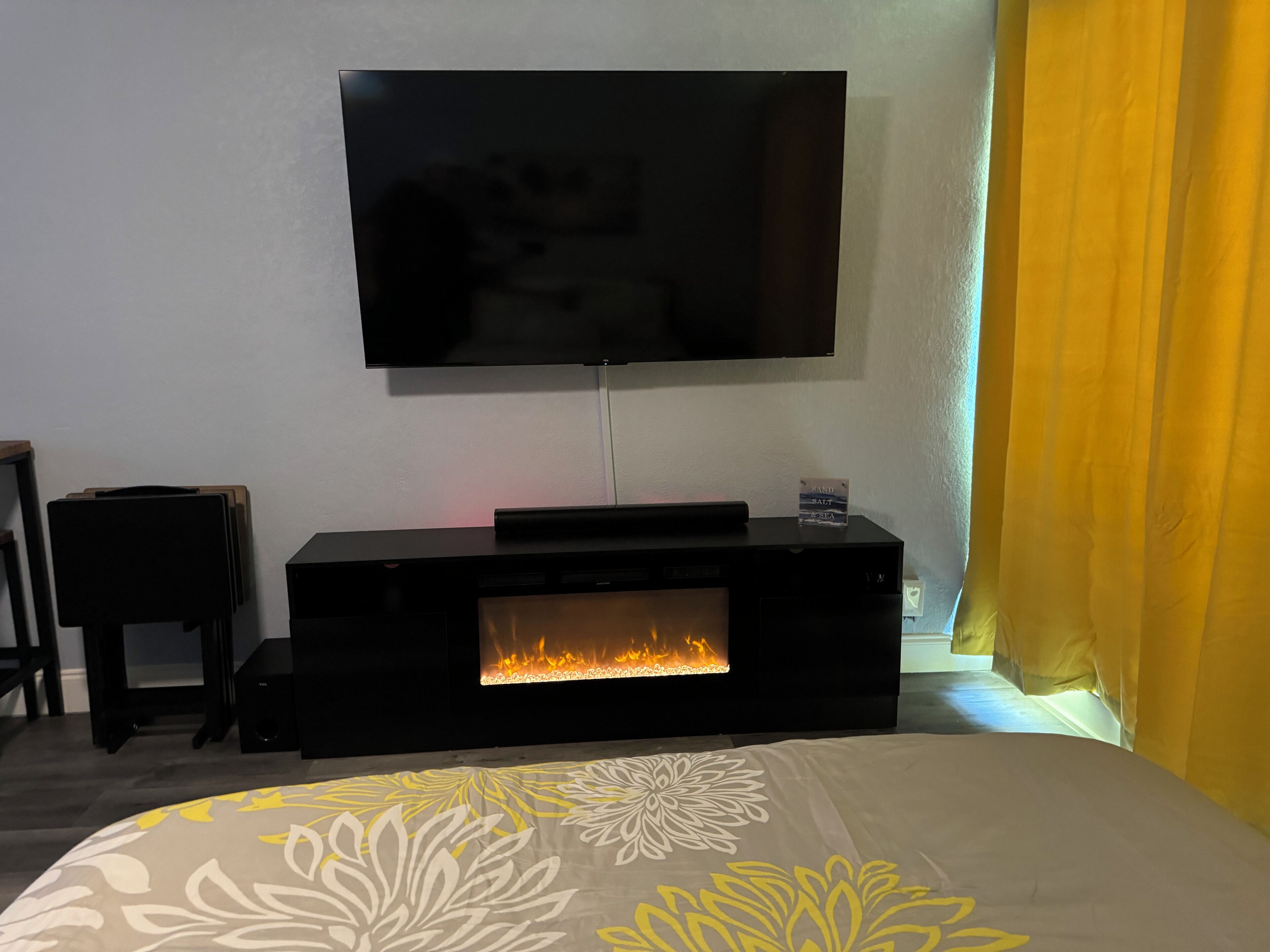 A room with a flat-screen TV mounted on the wall, a fireplace, and a bed with a floral-patterned bedspread.