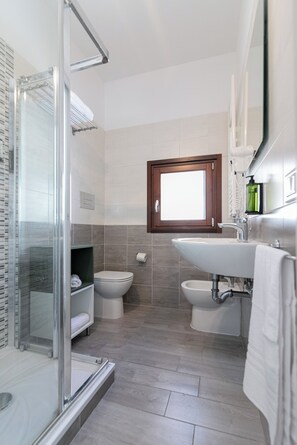 A modern bathroom with a glass shower enclosure, a toilet, a sink, and a window above the sink. - DeA ApartRooms (Termoli)