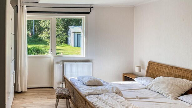 Une chambre à coucher comprenant un lit, une table de chevet, un fauteuil en osier et une fenêtre donnant sur un paysage verdoyant et un bâtiment situé à l’extérieur.