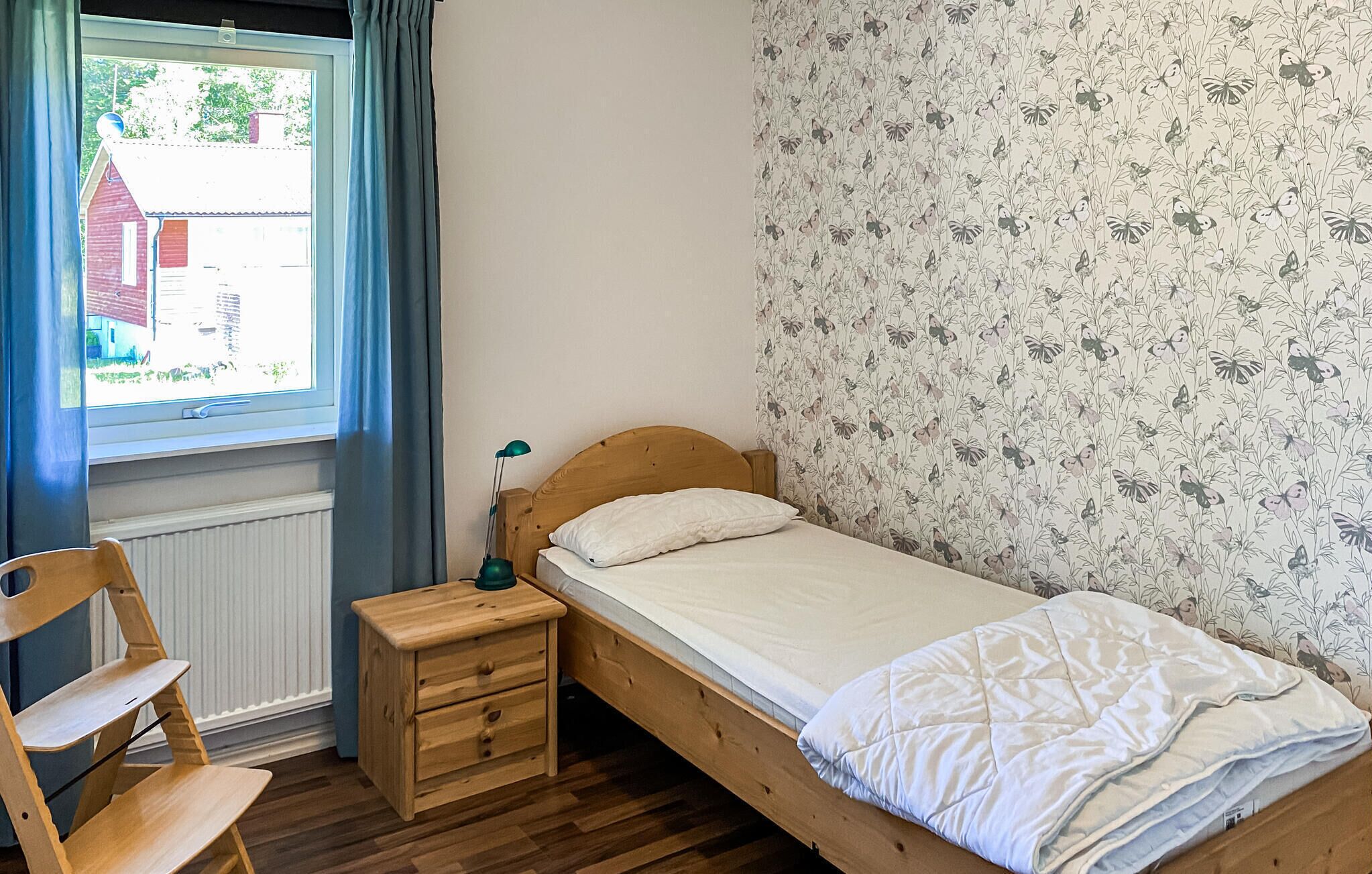 A small, single-bed room with a wooden headboard, a bedside table with a lamp, and a chair. A window with curtains is visible, and the wall has a floral wallpaper pattern.