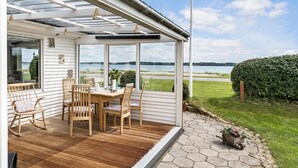 A covered patio with a wooden table and chairs, large windows offering a view of the lake, and a wooden wheelbarrow with flowers on the stone pathway.