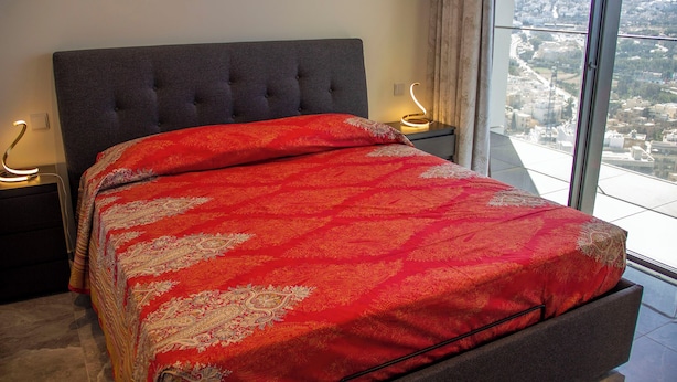 Un dormitorio con una cama grande, cabecera tapizada en gris y vistas a un paisaje urbano a través de un ventanal.