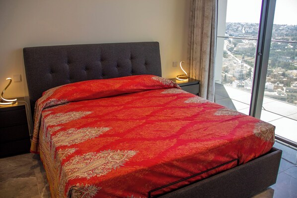 A bedroom with a large bed, grey upholstered headboard, and a view of a cityscape through a large window.