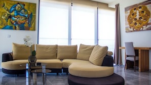Una sala de estar moderna con un sofá seccional color beige, una mesa de centro de vidrio y un jarrón con flores blancas. Hay dos ventanas grandes con cortinas translúcidas, y se puede ver un cuadro en la pared.