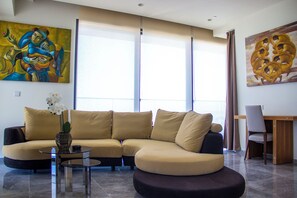 A modern living room with a beige sectional sofa, a glass coffee table, and a vase of white flowers. There are two large windows with sheer curtains, and a painting is visible on the wall.