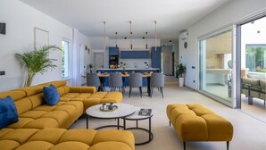 Un salon moderne avec un canapé, un pouf et une table basse. On aperçoit une cuisine à l’arrière-plan, avec des meubles bleus, ainsi qu’un coin repas avec des chaises.