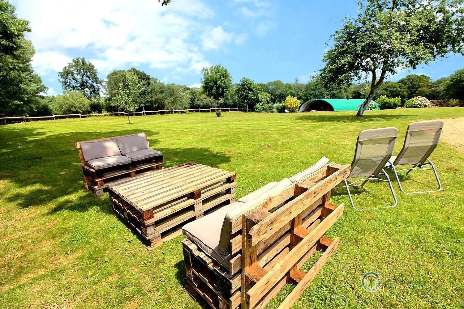 Un espace extérieur aménagé avec des bancs et des chaises en bois, une petite table et une tente verte, le tout installé dans un champ herbeux.