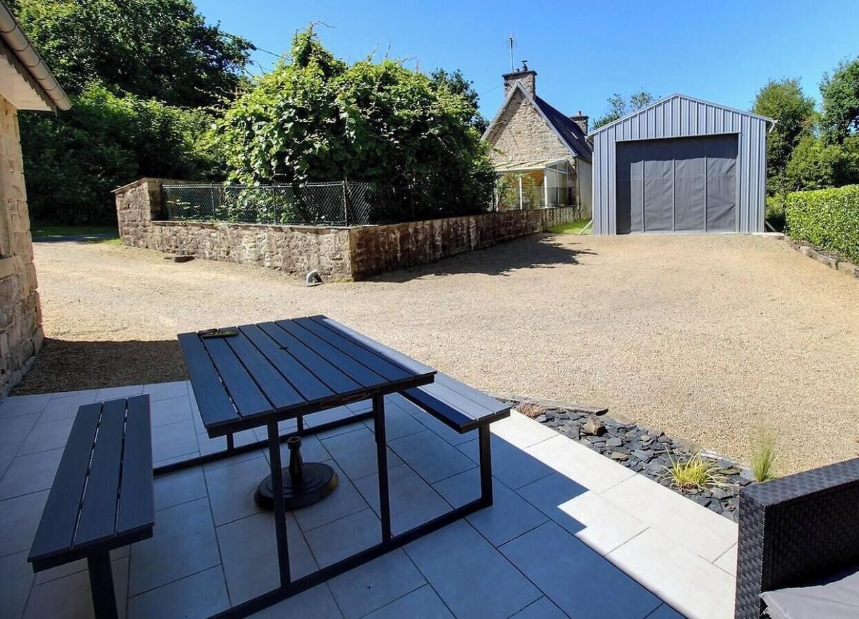 A patio with a table and benches, a stone wall, a shed, and a house in the background.