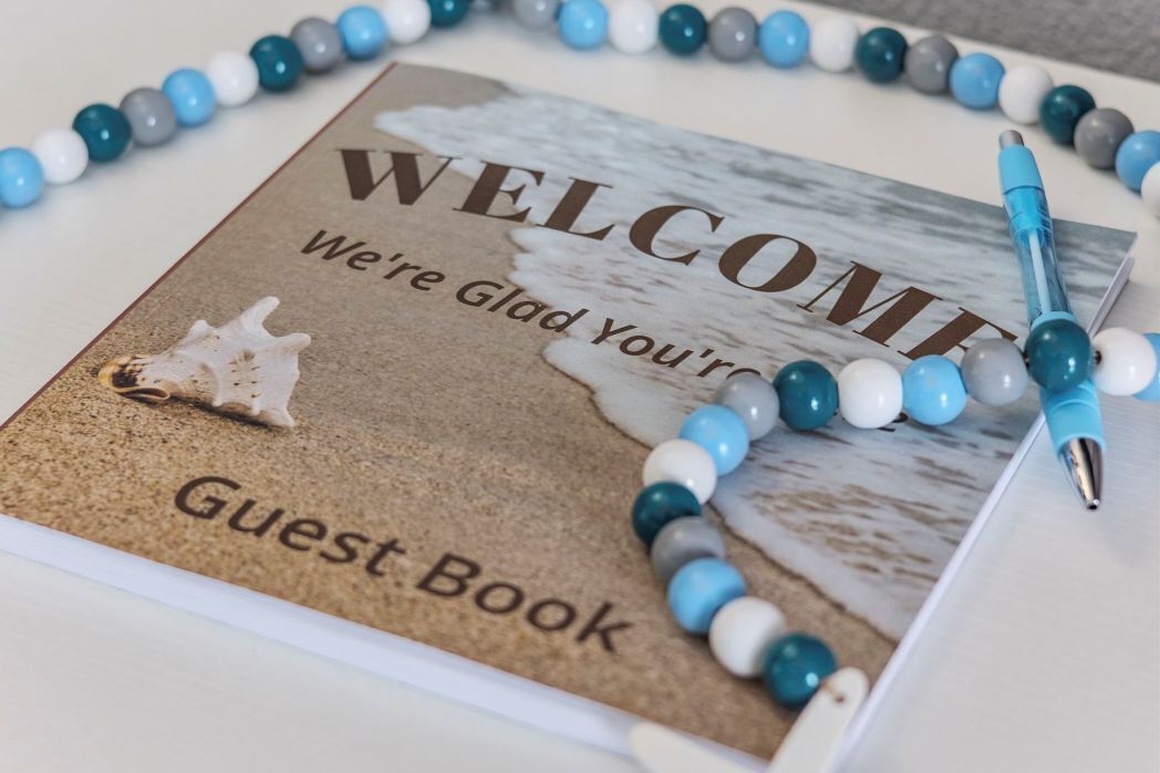A welcome sign for a guest book with a beach scene, a seashell, and a pen on a table.