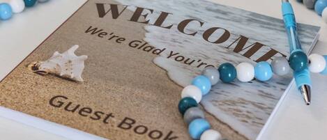 A welcome sign for a guest book with a beach scene, a seashell, and a pen on a table.