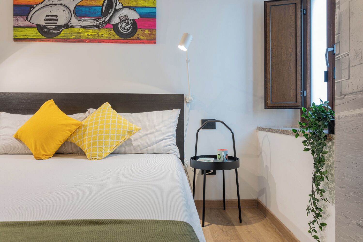 Une chambre à coucher avec un lit, deux oreillers, une table de chevet avec une tasse, une lampe sur pied, une plante et un tableau représentant un scooter.