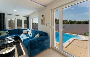 A modern living room with a blue sofa, a glass coffee table, and a view of a swimming pool through large sliding glass doors.