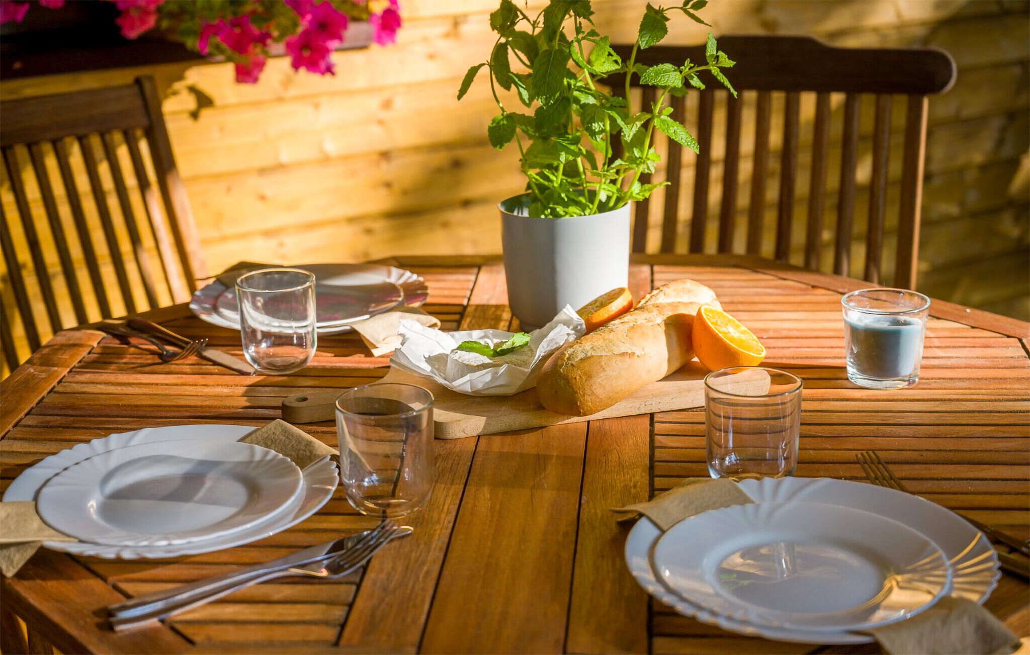 A wooden table set for a meal with plates, cutlery, glasses, a bread roll, an orange slice, and a potted plant.