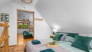 Un dormitorio abrigado y bien iluminado en el ático, con techo inclinado, piso de madera y una cama grande con ropa de cama verde y blanca. Se ve una escalera de madera y una pequeña mesa con una lámpara.