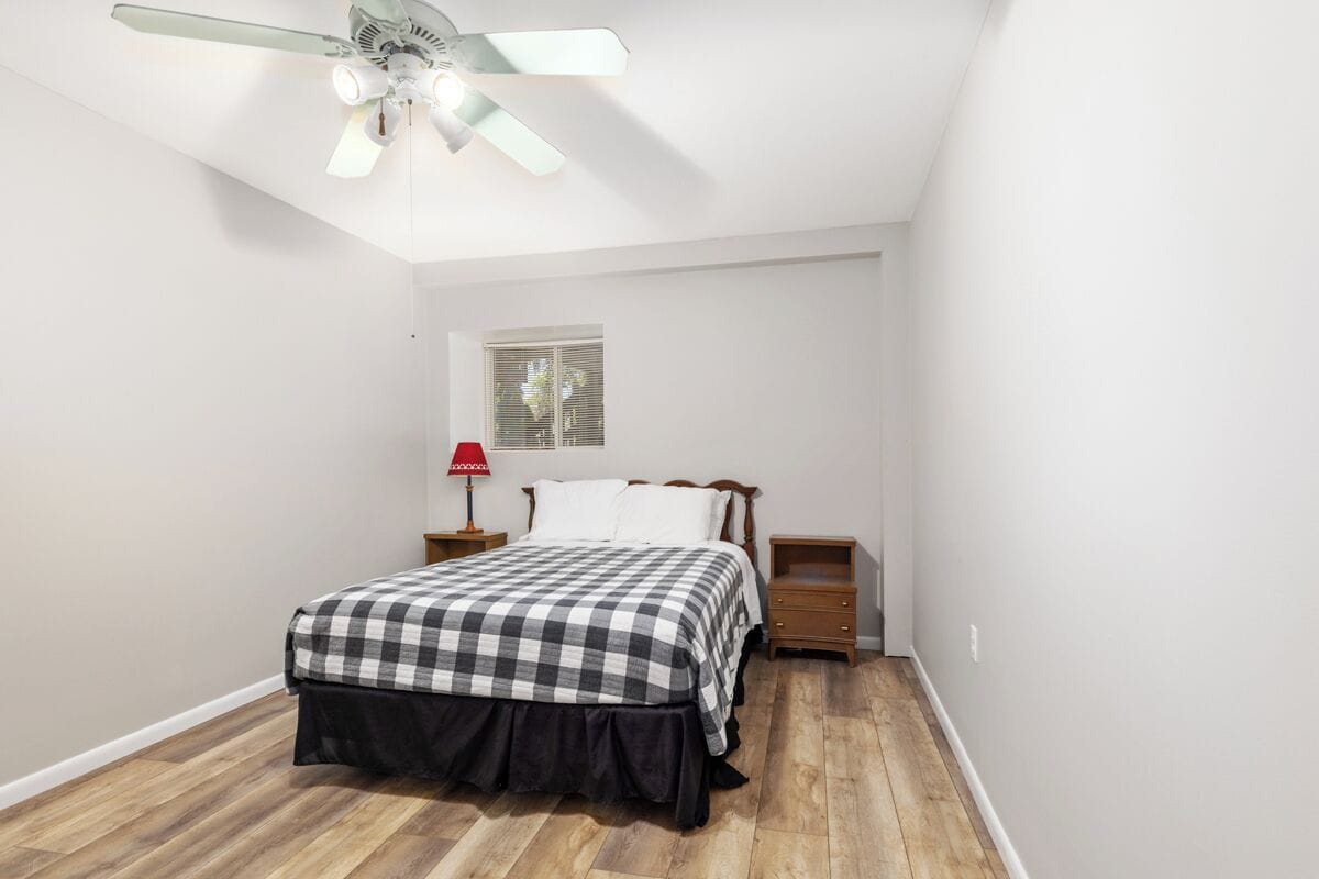 A bedroom with a bed, a nightstand, a lamp, a ceiling fan, and a window.