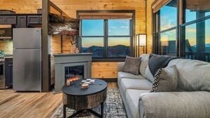 A cozy cabin interior with a fireplace, a sofa, and a view of the outdoors. - New Construction, Hot Tub, Mountain View, 4 Miles to Dollywood, Pac Man (Sevierville)