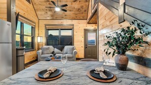A cozy cabin interior with a dining table set for two, a sofa, and a large window offering a view of the outdoors. - New Construction, Hot Tub, Mountain View, 4 Miles to Dollywood, Pac Man (Sevierville)