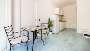 Une petite cuisine moderne avec des meubles blancs, un four encastré et un plan de travail. On y trouve un coin repas avec une table et des chaises, ainsi qu’une plante en pot.